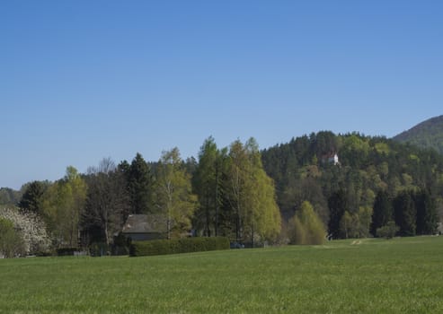 Spring landscape with view on village Marenice in Lusitian mountains with small chapel on hill, lush green grass meadow, deciduous and spruce tree forest and hills, blue sky background.