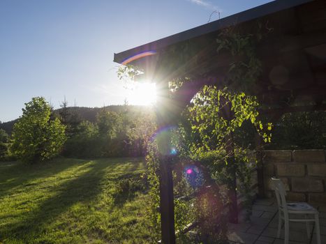 brown tiber wooden gazebo or pergola with climbing plants and flowers, chairs and surrounding sandstone wall, green grass blue sky in evening golden hour light, sun and lens flare