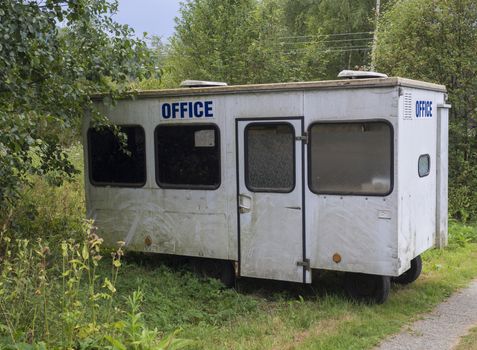 old small white trailer house with blue sign office, abandoned caravan in rural landscape near footpath, green grass and trees, blue sky