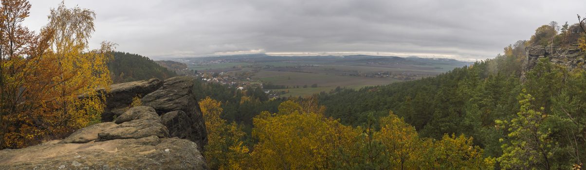 Panoramic view from sandstone pillars view-point at nature reserve Cesky Raj with autumn colored deciduous and coniferous tree forest and green hills, moody blue sky.