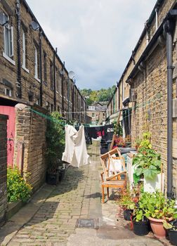 a back alley between streets with rows of traditional stone houses in hebden bridge west yorkshire with washing drying on lines and pot plants in bright summer sunshine
