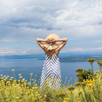 Rear view of young woman wearing striped summer dress and straw hat standing in super bloom of wildflowers, relaxing while enjoing beautiful view of Adriatic sea nature, Croatia.