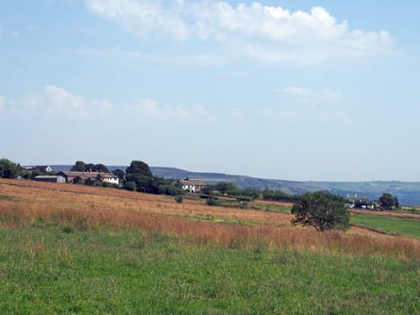 a panoramic view farmhouses between grass covered fields near blackshaw head in west yorkshire with distant pennine hills against a blue summer sky