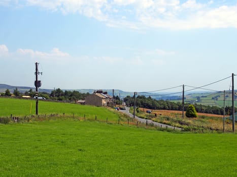 houses in the village of blackshaw head in west yorkshire with a road running thought fields with distant farmhouses and pennine hills against a blue summer sky