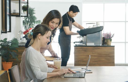 Beautiful young Caucasian women Using a laptop in the cafe. Asian female waitress serving espresso customers in a coffee shop. Background barista is preparing to make a drink according to order