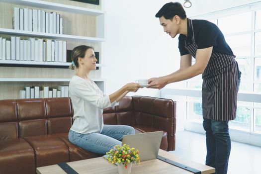 Beautiful young Caucasian women sitting, drinking coffee and using a laptop computer connected in the cafe. Asian barista young man doing hot espresso and service Served to customer according to order