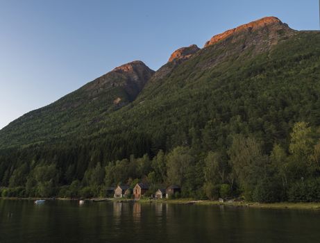 View of traditional wooden cottage along Hardanger Fjord in Kinsarvik in Norway with forest rocky hill, fishing boats and reflection in bay. Norwegian landscape, golden hour light and red enlighten cliff.