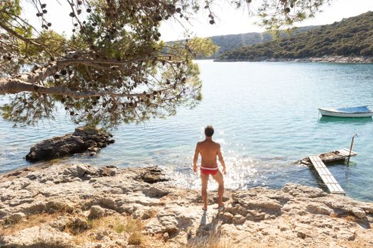 Rear view of man wearing red speedos tanning and realaxing on wild cove of Adriatic sea on a beach in shade of pine tree. Relaxed healthy lifestyle concept.