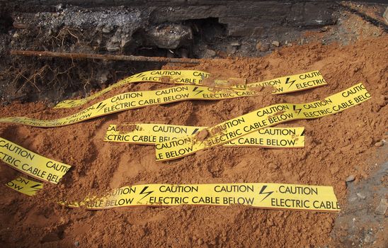 yellow tape signs warning of buried electric cable below in a trench being excavated during roadwork and building construction