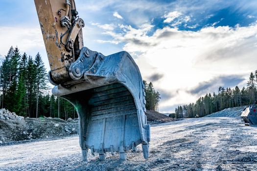 Close up back view at beautiful large excavator's scoop on new not yet ready road. Sunny evening with dark blue clouds, spring time, mixed forest with pile of big stones, dirt, sand at background.