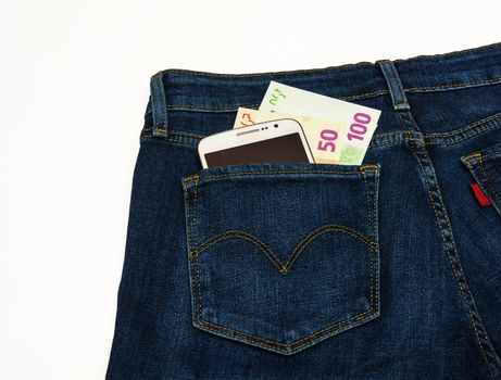 Trousers sewn from a blue denim lie on a white surface. In the back pocket of his pants is smart and banknotes.

