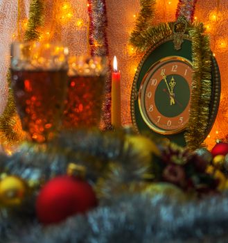 

In the foreground is m two glasses Christmas toys. Foreground out of focus. In the background, candle and clock. The clock time is five to twelve. unfocused visible spark and hours, in which time five minutes to twelve. Background illuminated garland.

