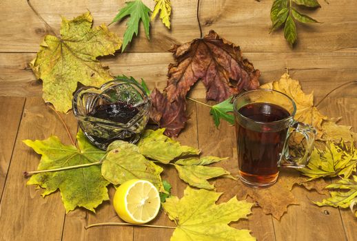 On a wooden surface lie autumn leaves, is a glass of tea and a bowl of jam and a number of lay sliced lemon