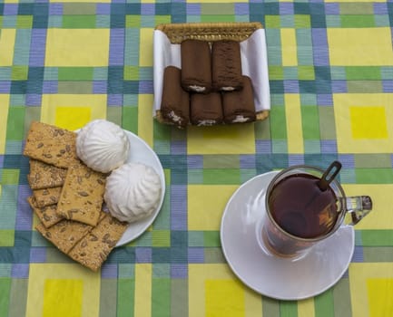 On a tablecloth in the drawing with squares is a cup of tea and biscuits with sunflower seeds and sesame seeds and chocolate rolls