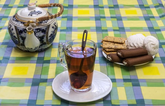 On the table mishandled colored tablecloth. On the tablecloth is a glass cup with tea, tea and sweets in the basket are