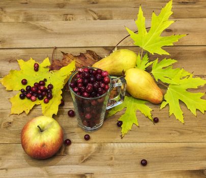 On a wooden surface is a glass beaker with cranberries. Next to them are autumn leaves, two pears and an apple.