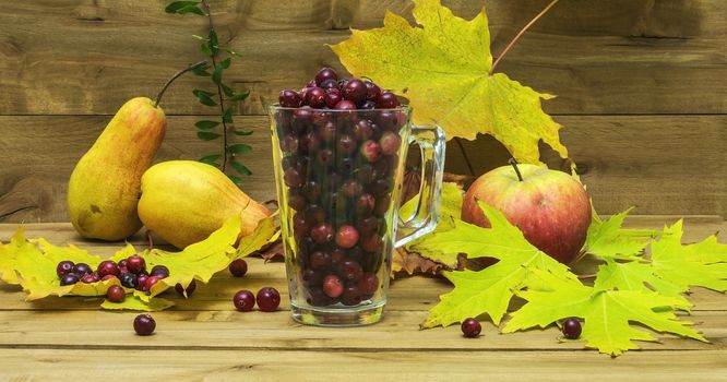 Against the background of the wooden surface is a glass beaker with cranberries, autumn leaves lie side by side, an apple and a pear.