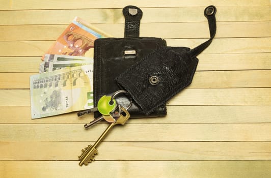 On a wooden surface lie the keys and unbuttoned the purse from which money can be seen euro banknotes