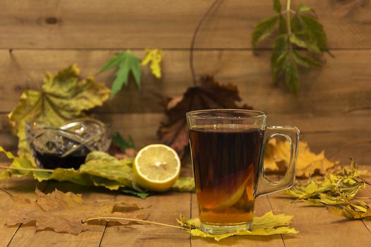 In the foreground is in focus glass cup of tea. In the background, the focus is visible without lemon, vase with jam and autumn leaves.