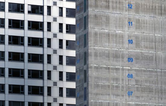the concrete core of a tall apartment building under construction with floor numbers marked in blue paint with silver cladding on a tall tower in the background