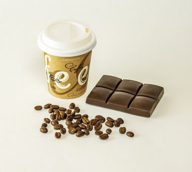 On a gray background is a paper cup of coffee, lying tiles of dark chocolate and coffee beans scattered