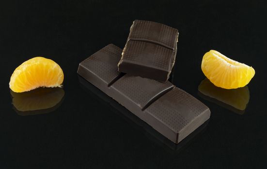 On a dark background are a mirror reflection of a bar of chocolate and slices of mandarin