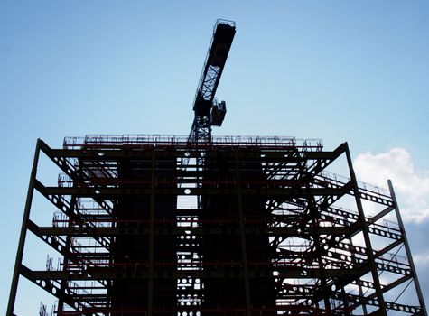 a large construction site in silhouette in the evening with steel girders and a crane in silhouette against a blue cloudy sky