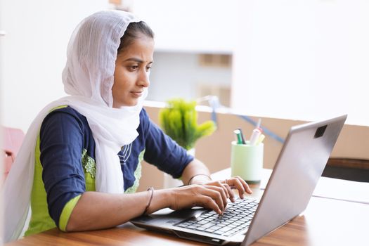 Happy young indian girl using computer looking at screen working in internet - young girl busy working or reading on laptop at home - concept of online education, work from home or wfh.