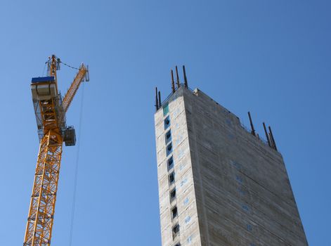 a yellow tower crane working on a construction site with a tall concrete tower against a bright blue sky