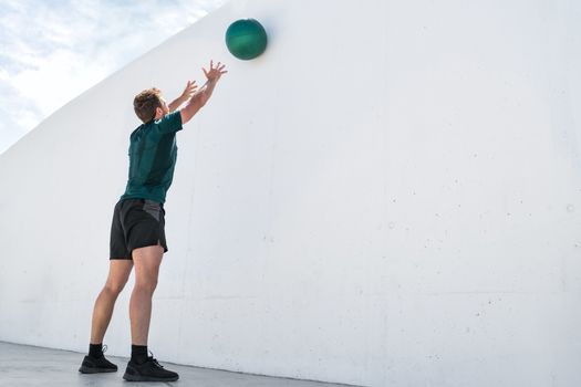 Strength training fit man cross training throwing medicine ball on gym wall at fitness centre. Workout bodyweight exercises using heavy weight ball. Overhead medicine ball throw plyometric exercise.
