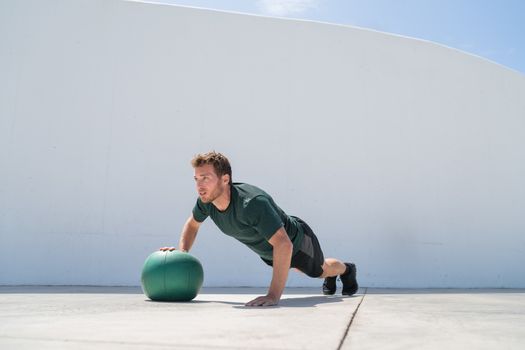 Fitness man strength training pushup chest and shoulder muscles doing alternating single arm medicine ball push-ups floor exercises at outdoor gym.