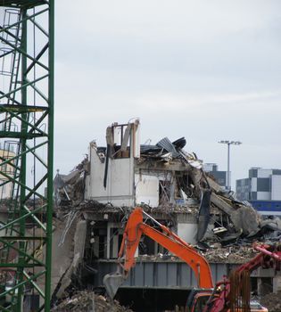 an orange digger with a backhoe working on a large urban demolition site with a metal tower crane at the edge of the frame and grey sky