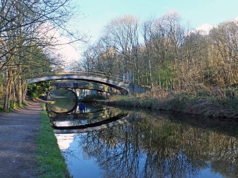 two bridges crossing the leeds to liverpool canal in armley with trees reflected on the water and footpath