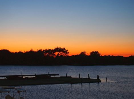 twilight over a lake with the sun setting behind a tree covered island with glowing orange sky and a jetty in silhouette