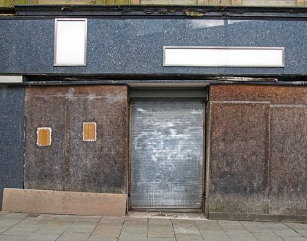 a street view of the front of an old abandoned shop with decaying boarded up windows covering the storefront and a closed steel shutter