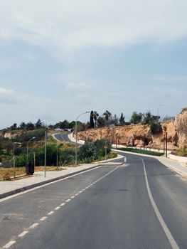 an empty two lane road with pavements and lampposts curving up a hill into the distance with surrounding trees and blue sunlit sky