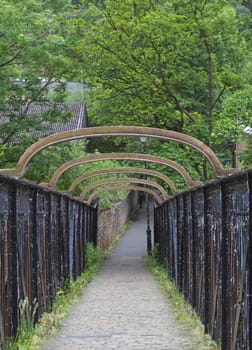 an old metal pedestrian footbridge crossing a railway line leading to a narrow alley surrounded by trees and rustic buildings