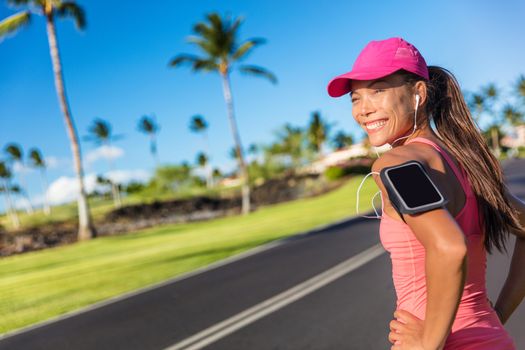 Fitness motivation running music woman runner. Happy jogging girl listening to smartphone app with earphones. Asian athlete wearing cap and armband smart phone holder outdoor on city road.