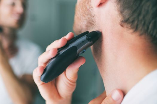 Man shaving beard using electric trimmer shaver. Male beauty grooming concept. Home lifestyle young person looking at bathroom mirror trimming hair on neck.