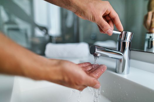 Man washing hands in bathroom sink at home checking temperature touching running water with hand. Closeup on fingers under hot water out of a faucet of a sink.