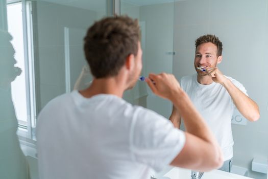 Man getting ready in the morning doing hygiene routine brushing his teeth looking in mirror of home bathroom, using toothbrush in for clean dental oral care.