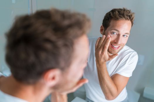 Man putting skincare facial treatment cream on face. Anti-aging skin care product. Male beauty morning routine at home lifestyle. Guy looking in bathroom mirror applying moisturizer under eyes.