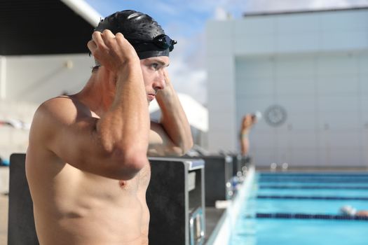 Swimmer man athlete getting ready for swim exercise at outdoor swimming pool sport putting on cap and goggles at training center.