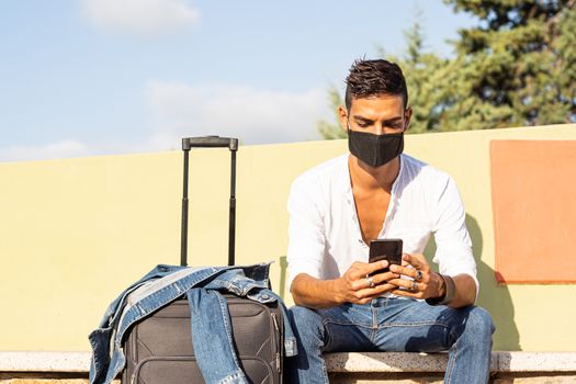 Young stylish caucasian man traveler with a black COVID protection mask using the smartphone sitting on a colorful bench with the denim jacket on a traveling bag - Coronavirus safe traveling concept