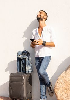 Happy young stylish caucasian man traveler smiling using the smartphone leaning against a white wall with traveling bag on the ground - Alwais connected concept - Solo male millenial traveller