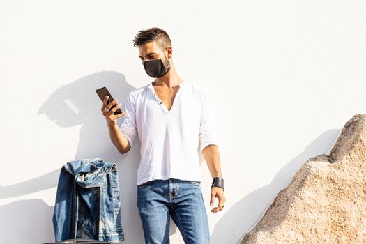 Stylish millennial boy standing to a white wall with COVID protection mask using his smartphone - Solo traveller safe concept - Connected young man communicating with technology