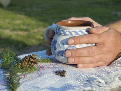 Close up hand of male holding hot smoking tea in rustic white blue painted ceramic cup, white tablecloth, green pine branches, cone and cloves spices, green background, golden light. Winter time coming concept.