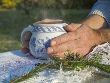 Close up hand of male holding hot smoking tea in rustic white blue painted ceramic cup, white tablecloth, green pine branches, cone and cloves spices, green background, golden light. Winter time coming concept.