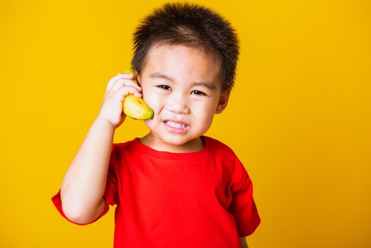 Happy portrait Asian child or kid cute little boy attractive smile wearing red t-shirt playing holds banana fruit pretending to be like a telephone, studio shot isolated on yellow background