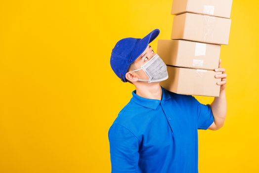 Asian young delivery worker man in blue t-shirt and cap uniform wearing face mask protective lifting stack a lot of boxes, under coronavirus or COVID-19, studio shot isolated yellow background